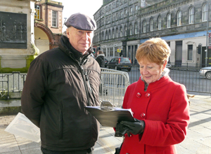 Malcolm in winter wear with a woman in a red coat signing a petition beside the Queen Victoria statue